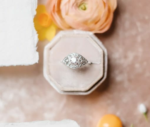 A fine diamond ring in a fancy ring box being shown from an upward angle, laid on a table with flowers and white cloth.