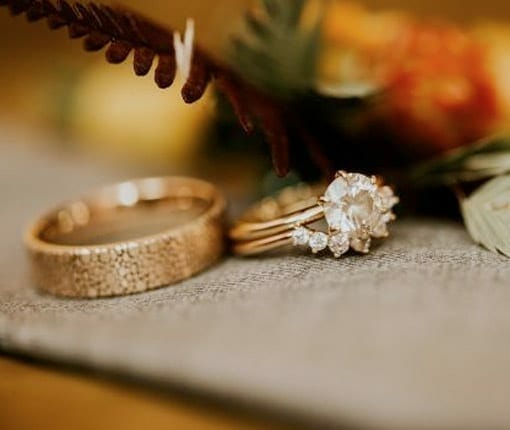 A gold wedding band and a gold engagement and wedding band set beside each other and laid on a wooden table with flowers and a cloth.