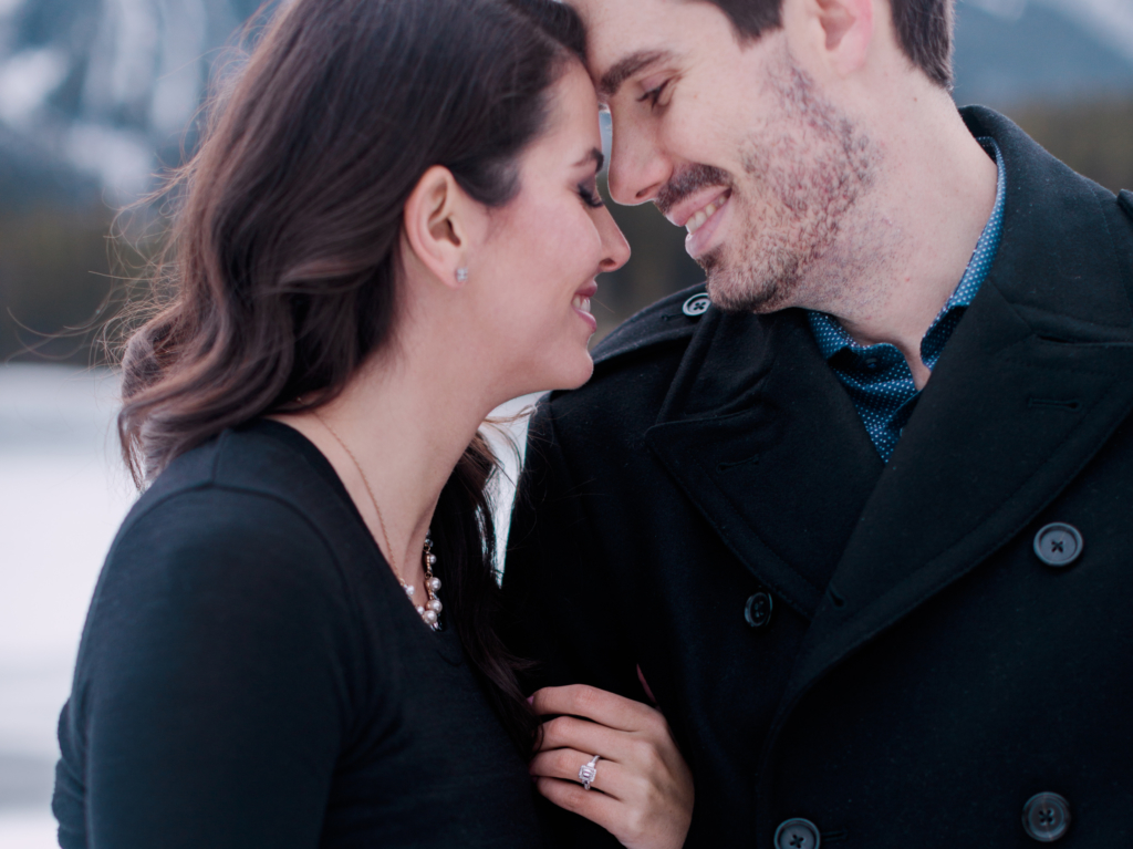 A newly engaged couple dressed in blakc winter clothing posing by hold each other close for their engagement photos.