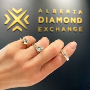 Someone holding three diamond engagement rings with different settings in front of the ADX logo.